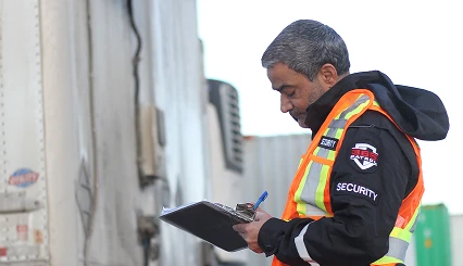 A security officer checking shipping documents on a clipboard next to a semi-truck, performing logistics Calgary security service duties.